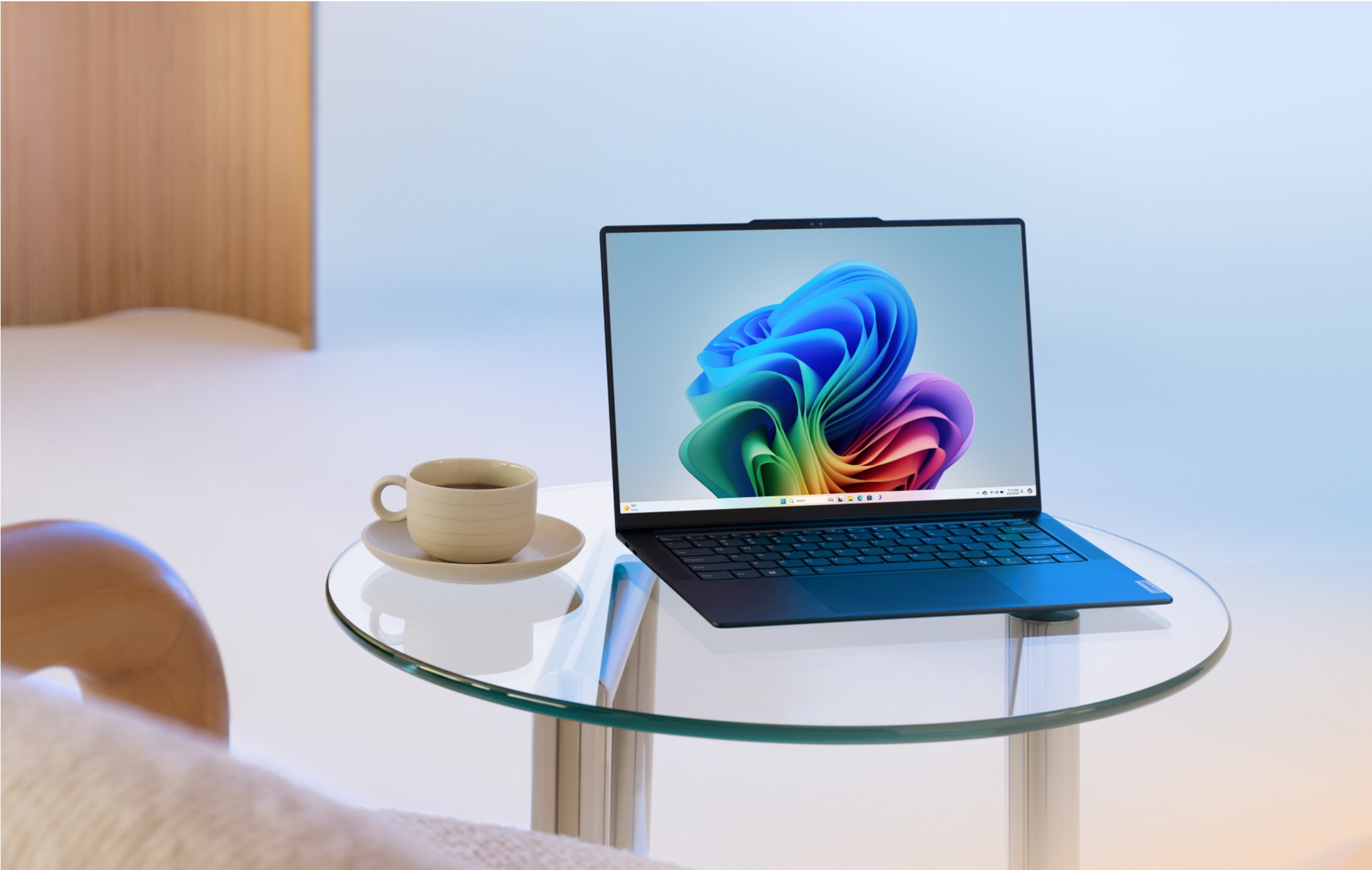 Ein Windows 11-PC mit mehrfarbigem Bloom-Desktophintergrund. Der Laptop befindet sich auf einem kleinen, kreisförmigen Glastisch neben einer Kaffeetasse.